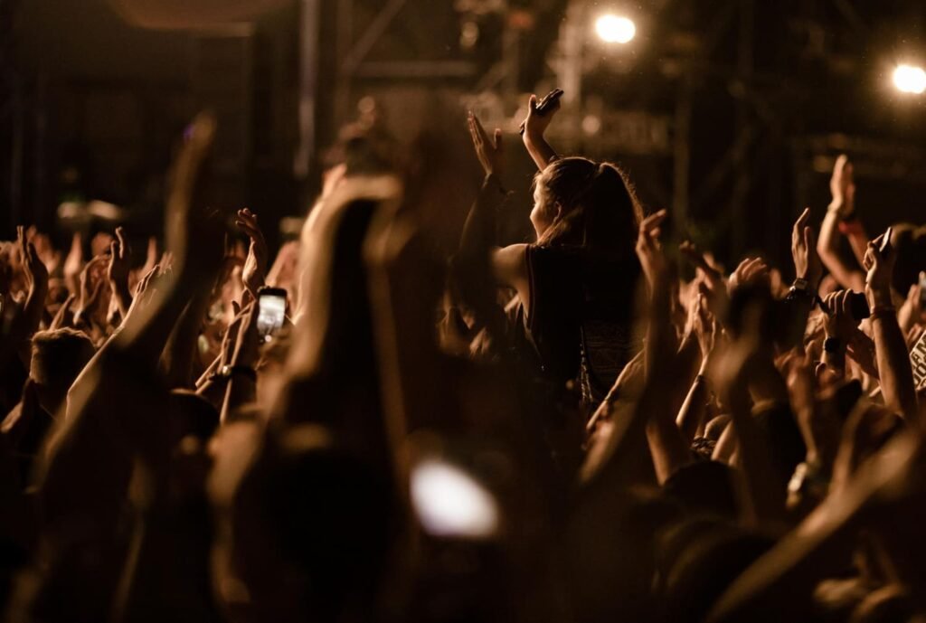 Multitud de personas disfrutando de un concierto, aplaudiendo y levantando las manos bajo una iluminación cálida y luces de escenario en el fondo Vida Nocturna en Querétaro