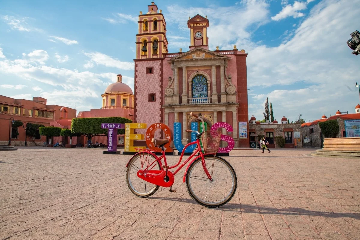 Vista de la iglesia de Santa María de la Asunción en Tequisquiapan, Querétaro, con una bicicleta roja en primer plano y las letras coloridas de Tequisquiapan al fondo.
