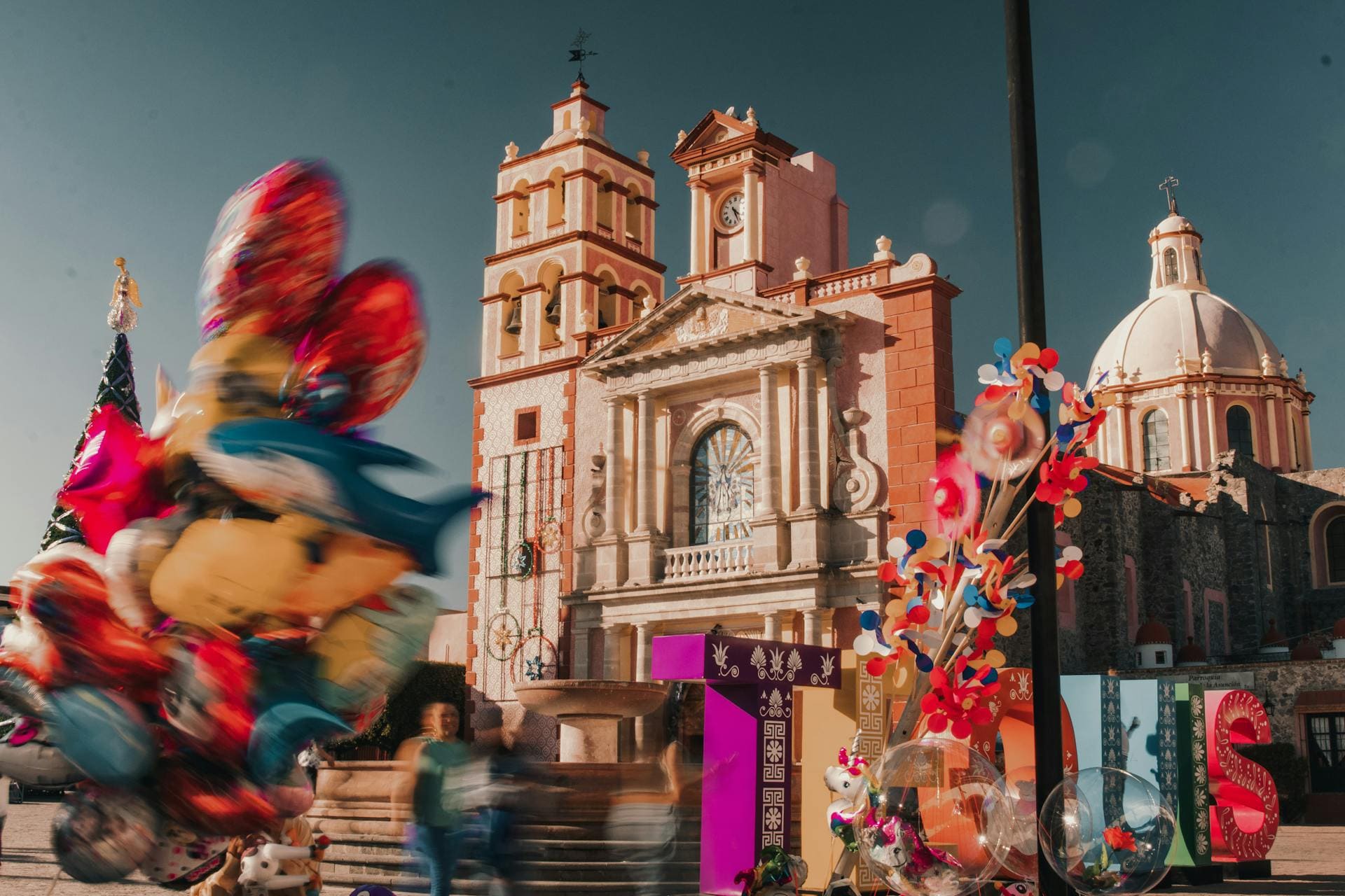 Vista de la iglesia neoclásica de Santa María de la Asunción en Tequisquiapan, Querétaro, con un vendedor de globos coloridos en primer plano.