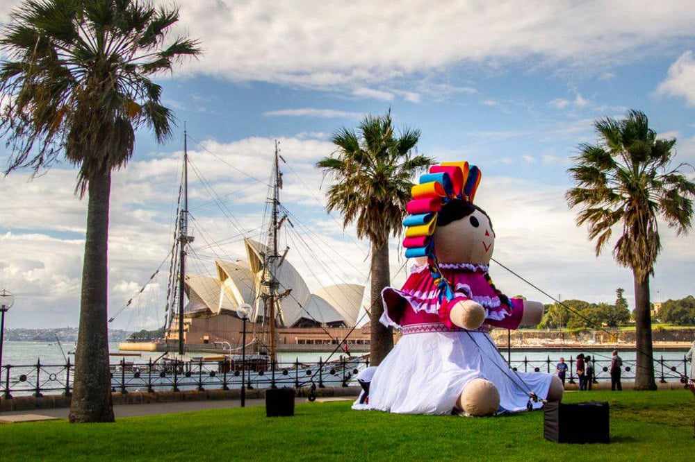 Muñeca Lele Gigante en Sídney: Símbolo Cultural de México en el Mundo Muñeca Lele gigante con vestido tradicional otomí en Sídney, Australia, con la Ópera de Sídney y un velero de fondo.