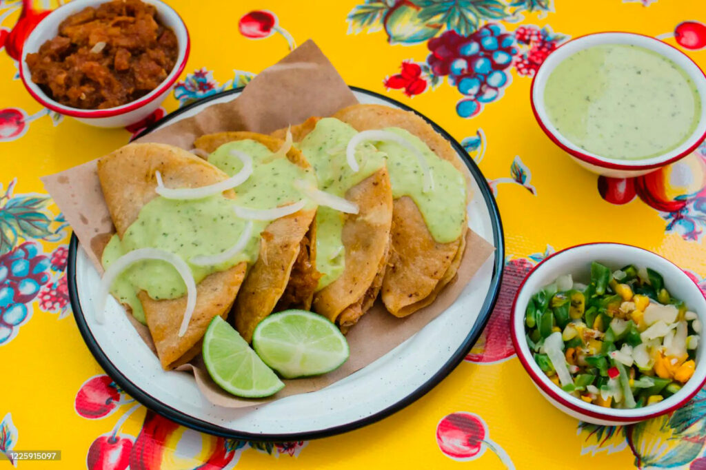 Tres Tacos de Canasta con Salsa Verde en Plato Rústico Tres apetitosos tacos de canasta apilados en un plato de cerámica verde texturizado, acompañados de un pequeño recipiente cuadrado con salsa verde brillante. La luz resalta la textura de los tacos y la salsa.