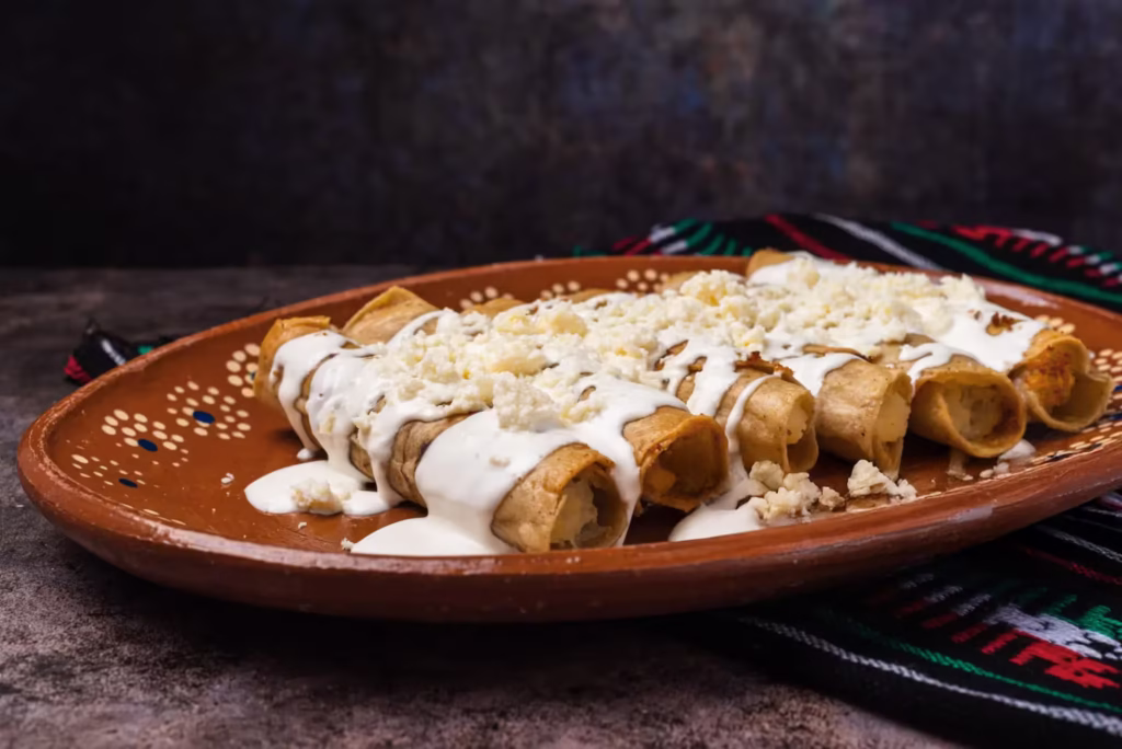 Tacos dorados en plato de barro | Tradición servida con crema y sabor auténtico Seis tacos dorados mexicanos en plato de barro, cubiertos con crema y queso blanco rallado.