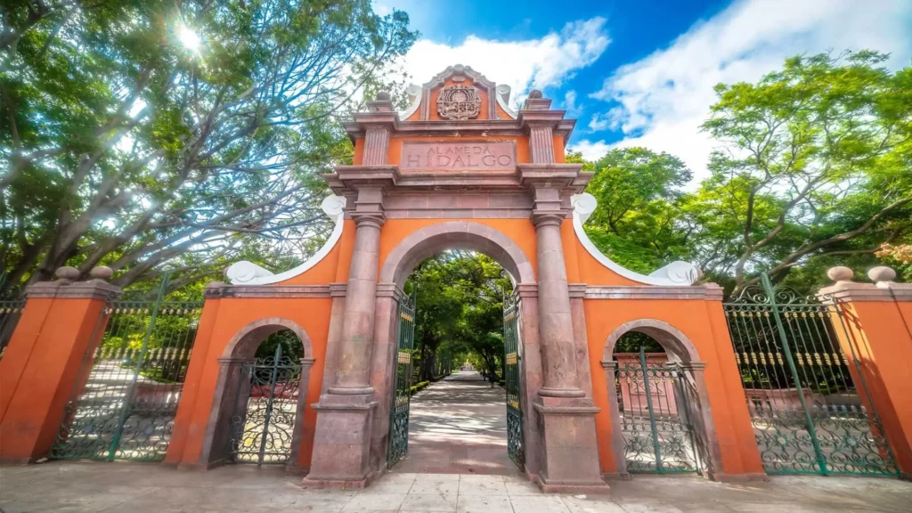 Acceso lateral de la Alameda Hidalgo en Querétaro con arco de cantera, rejas verdes y arbolado urbano.