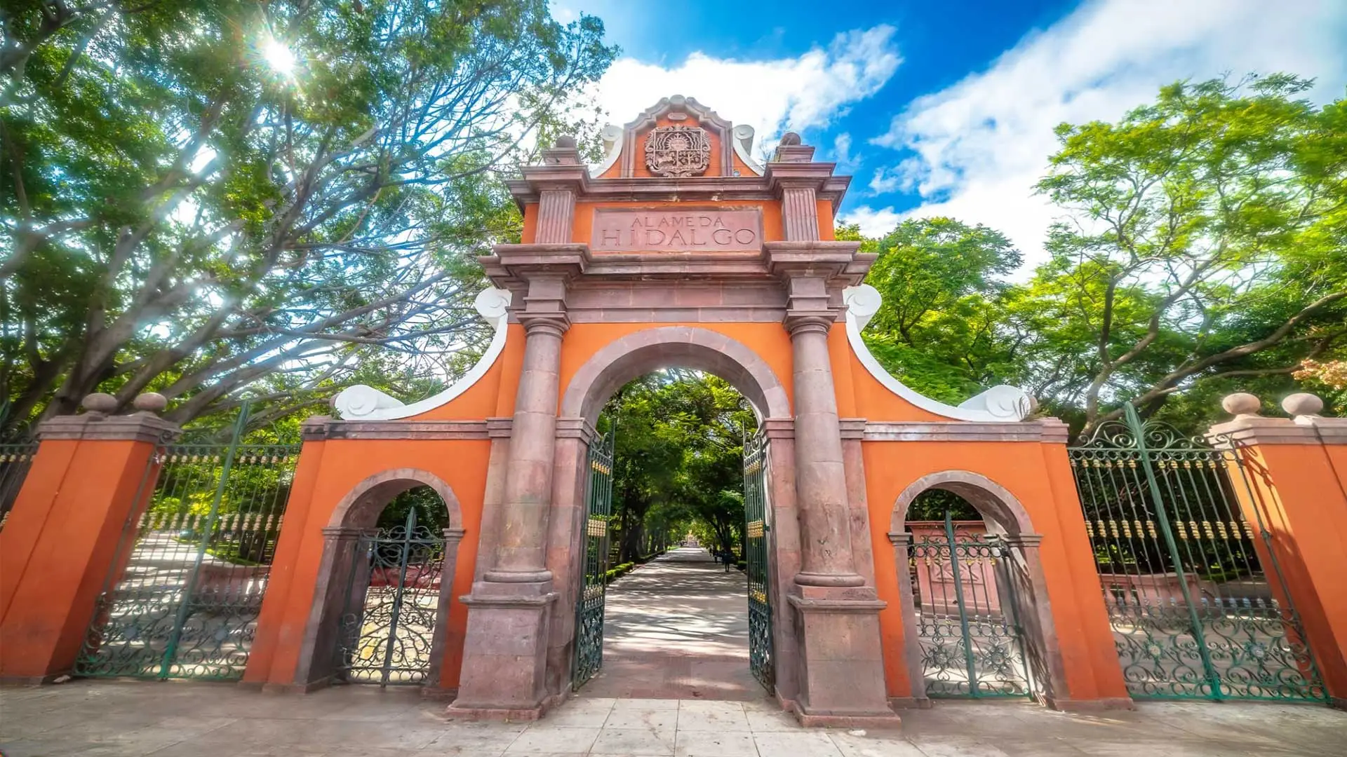 Acceso lateral de la Alameda Hidalgo en Querétaro con arco de cantera, rejas verdes y arbolado urbano.