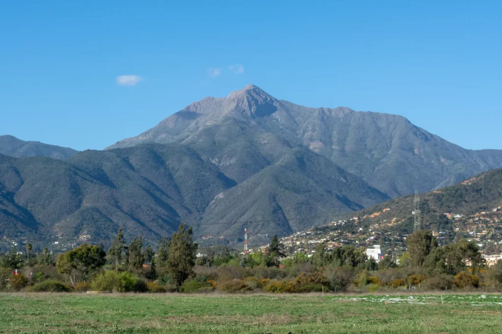 Montaña de cima rocosa con viviendas al pie y un campo en primer plano.