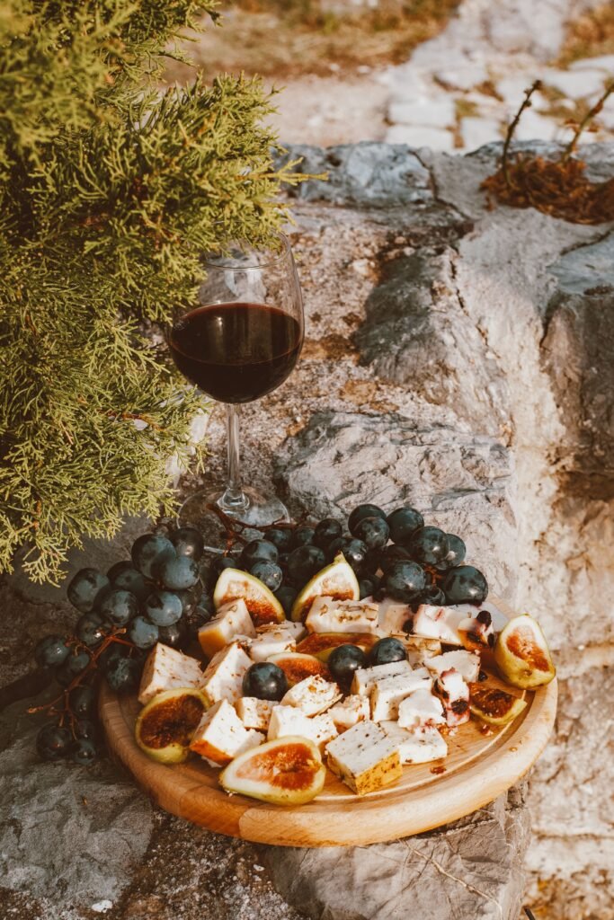 Imagen ilustrativa de una tabla de queso, fruta y una copa de vino al aire libre